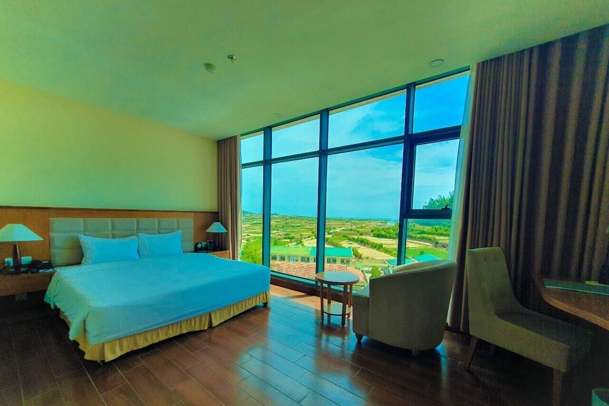 Ein Hotelzimmer mit einem großen Bett, einem Sessel, einem kleinen Tisch und Blick auf eine Landschaft durch große Fenster.