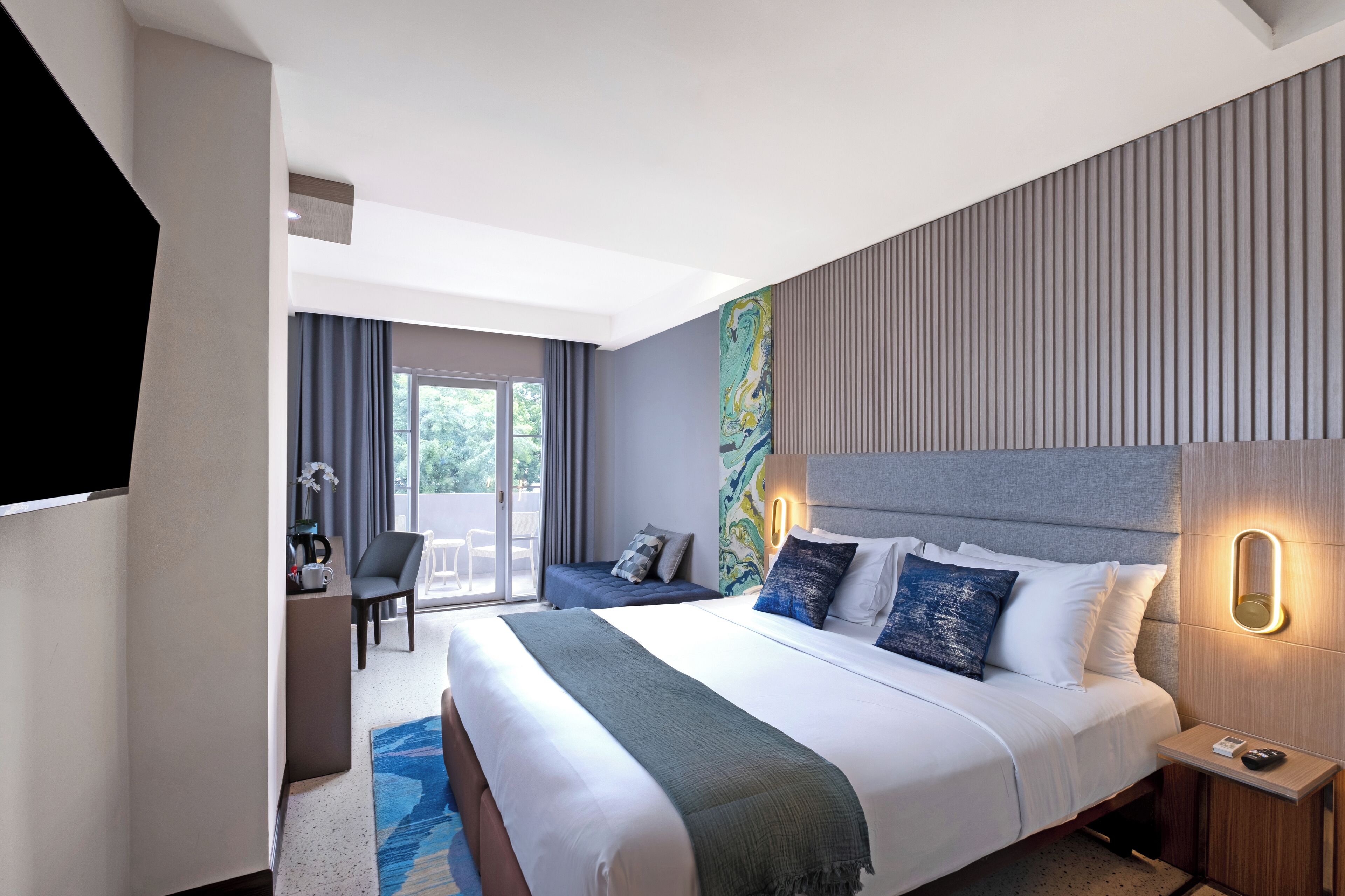A modern hotel room with a large bed, a desk with a chair, and a view of the outdoors through a sliding door.