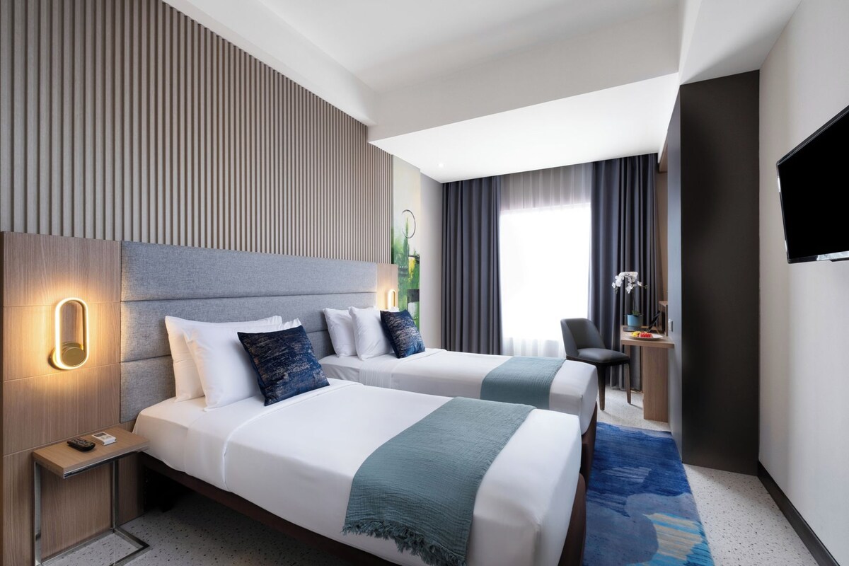 A hotel room with two beds, a desk, a TV, and a window with curtains.