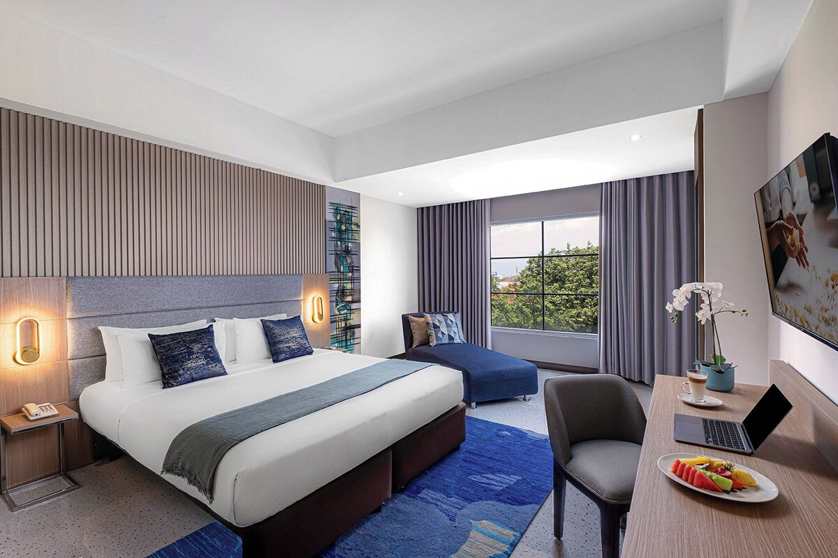 A modern hotel room with a large bed, a desk with a laptop, a chair, a small table with a drink, and a window with a view.