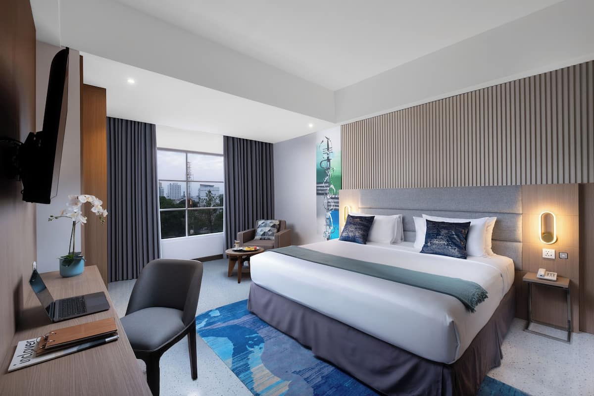 Ett modernt hotellrum med en stor säng, ett skrivbord med en bärbar dator, en stol, ett litet bord och utsikt över staden genom fönstret.
