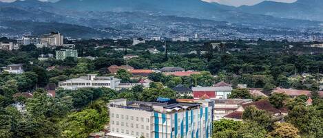 A cityscape with a prominent building featuring a modern design with a blue and white facade, surrounded by dense greenery and a backdrop of mountains.