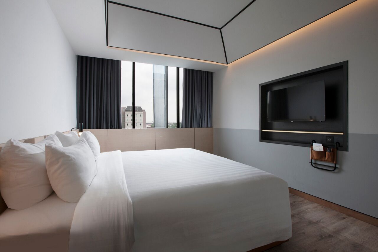 A modern hotel room with a large bed, a flat-screen TV mounted on the wall, and a city view through the window.