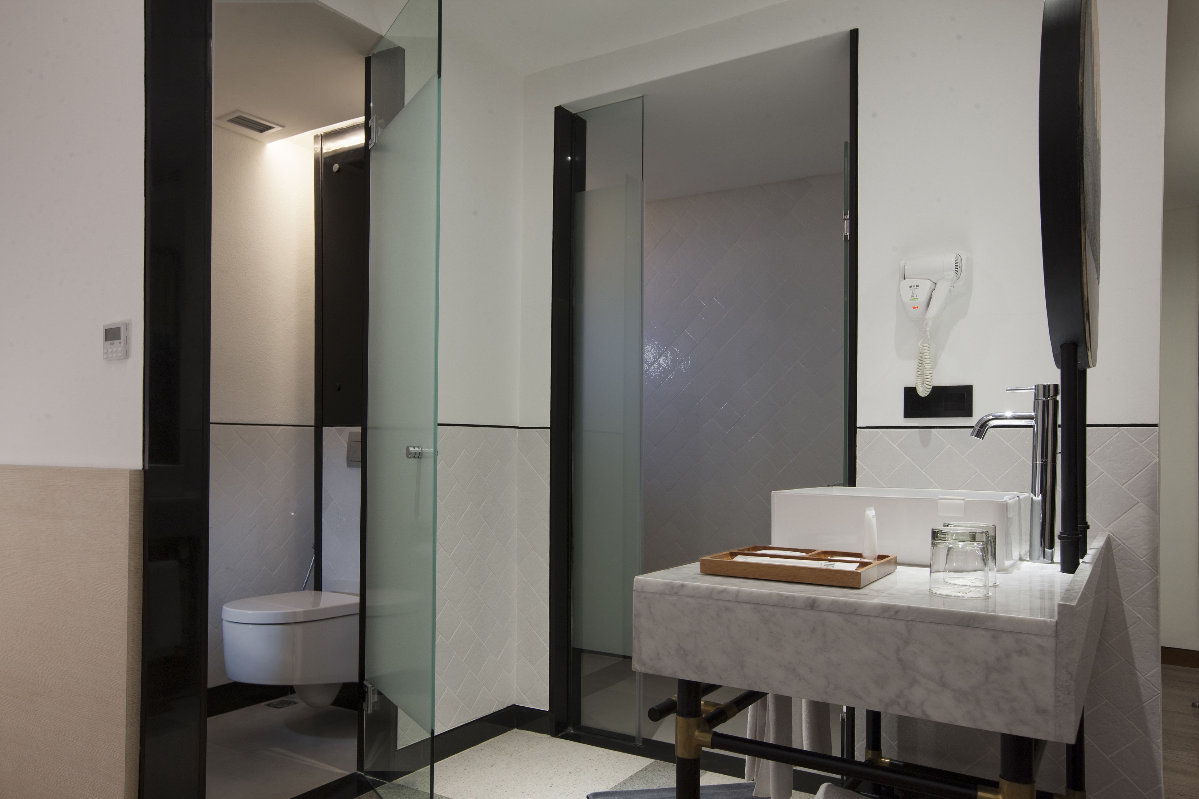 Un baño moderno con cabina de ducha de vidrio, un lavabo blanco con encimera de mármol y un secador de pelo fijado a la pared.
