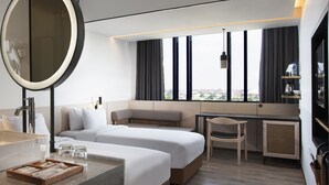 Habitación de hotel moderna con una cama grande, un escritorio, una silla, un espejo redondo y vistas de la ciudad a través de amplias ventanas.