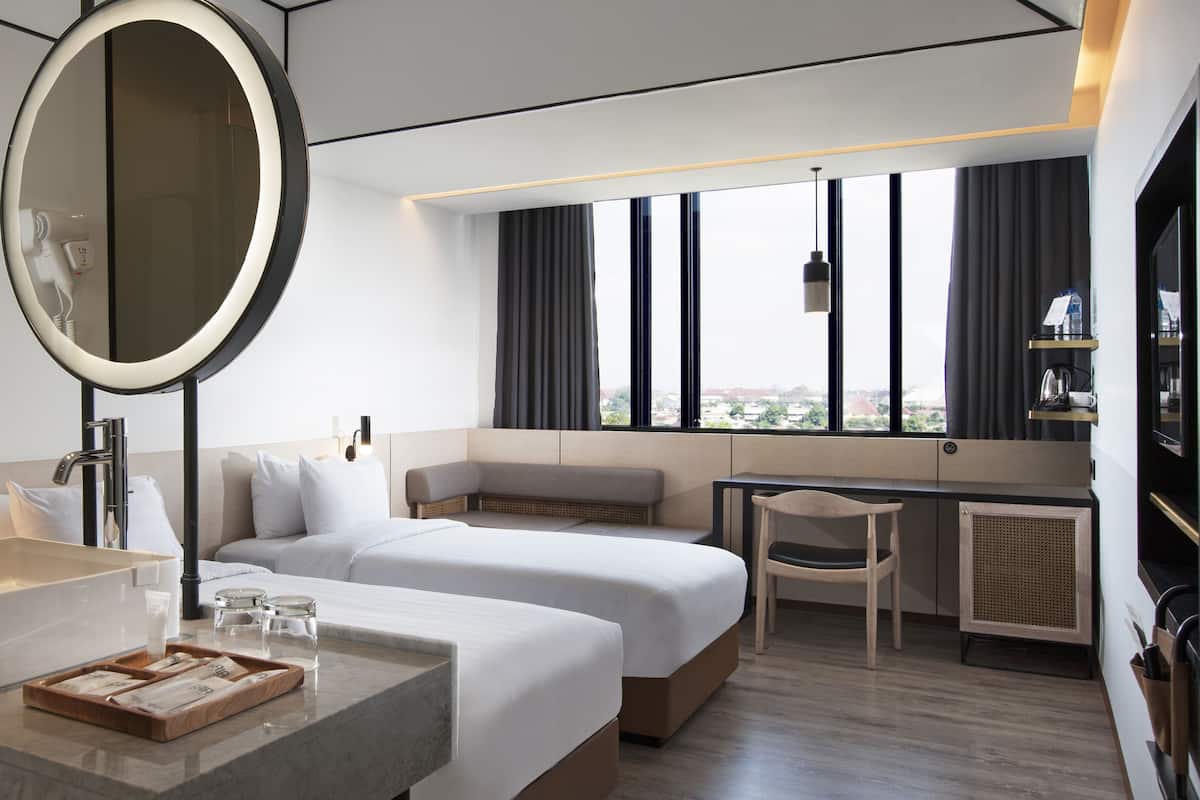 A modern hotel room with a large bed, a desk, a chair, a round mirror, and a view of the city through large windows.