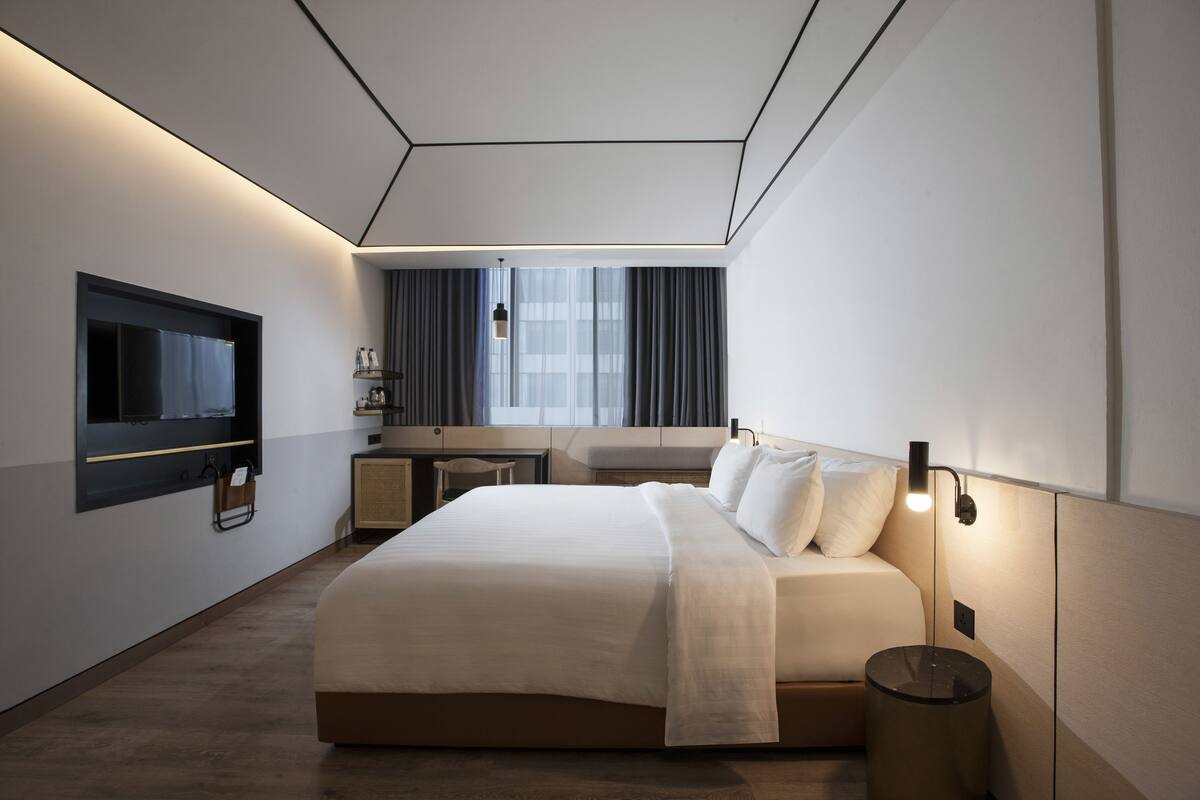 A modern hotel room with a large bed, a flat-screen TV mounted on the wall, a bedside table, and a small desk with a chair.