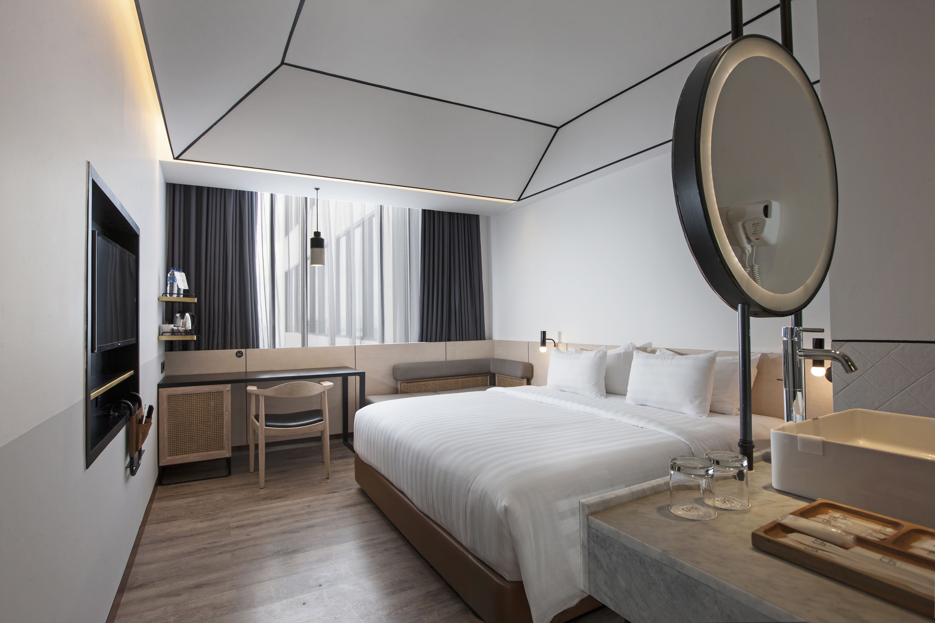 Une chambre d’hôtel moderne dotée d’un grand lit, d’un bureau avec une chaise, d’un petit coin salon, d’un grand miroir et d’une salle de bain avec un lavabo et un miroir.