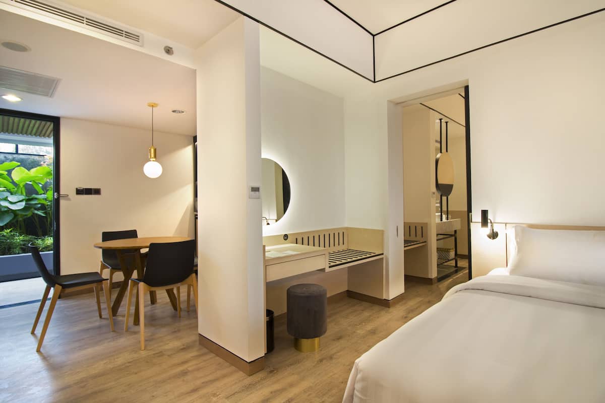 A modern hotel room with a wooden floor, a bed, a desk with chairs, a mirror, and a small stool.