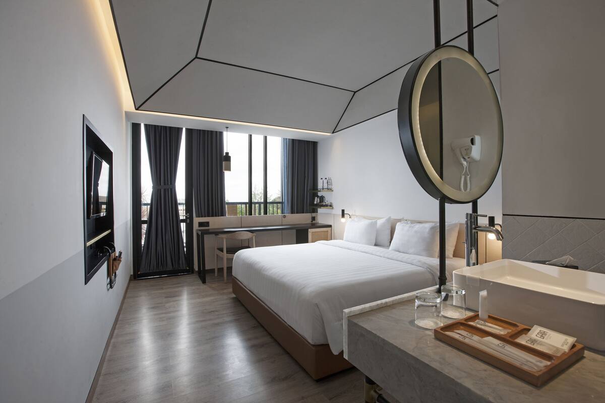 A modern hotel room with a large bed, a desk, a bathroom with a large mirror, and a view of the outdoors.