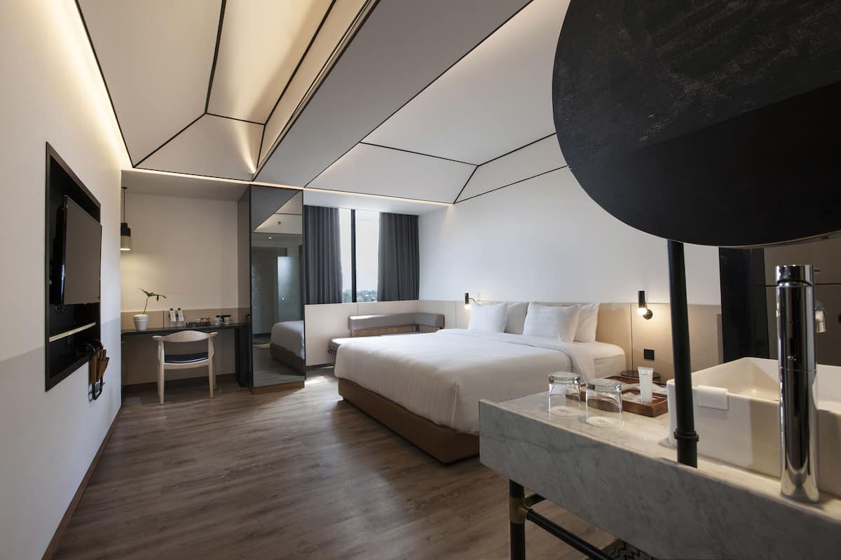 A modern hotel room with a large bed, a desk, and a bathroom with a mirror and sink.