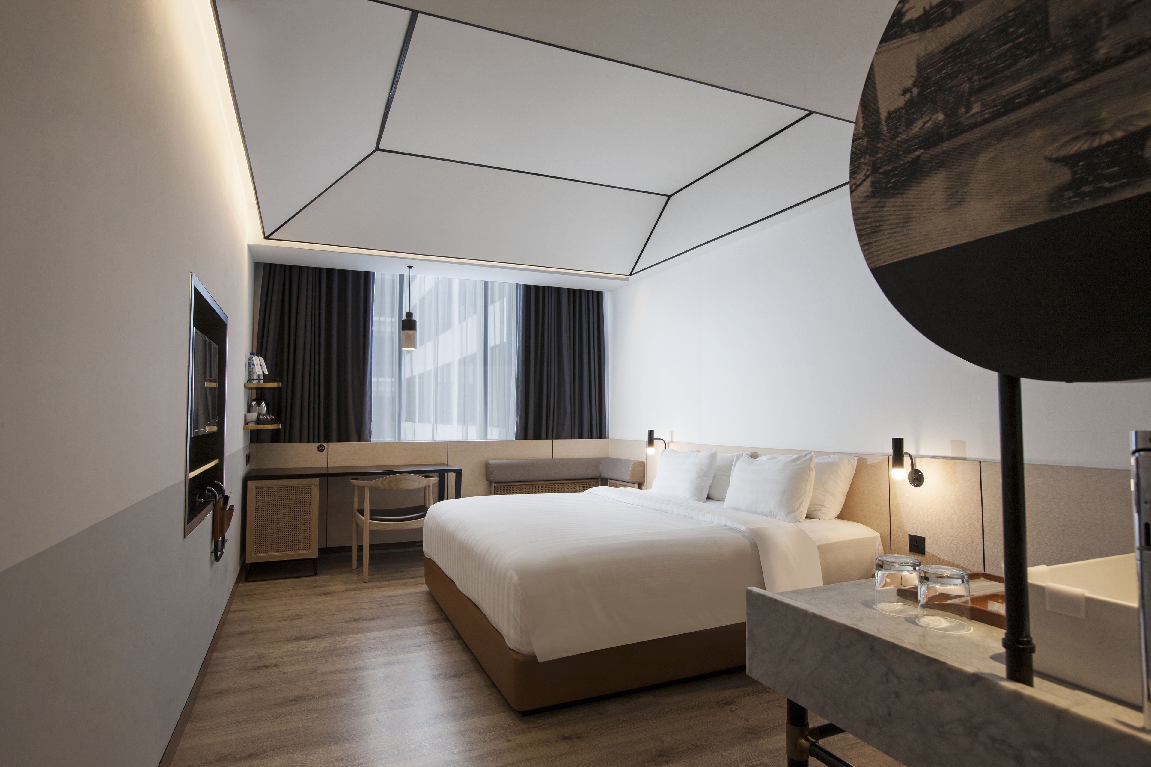 Una habitación de hotel moderna con una cama grande, un escritorio, una silla, un tocador con espejo y un amplio ventanal con cortinas.