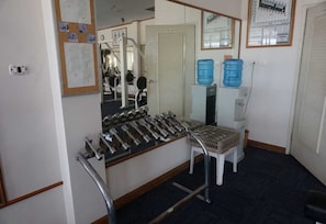 Una habitación con una banca de pesas, un espejo, un dispensador de agua y una puerta con un tablón de anuncios.