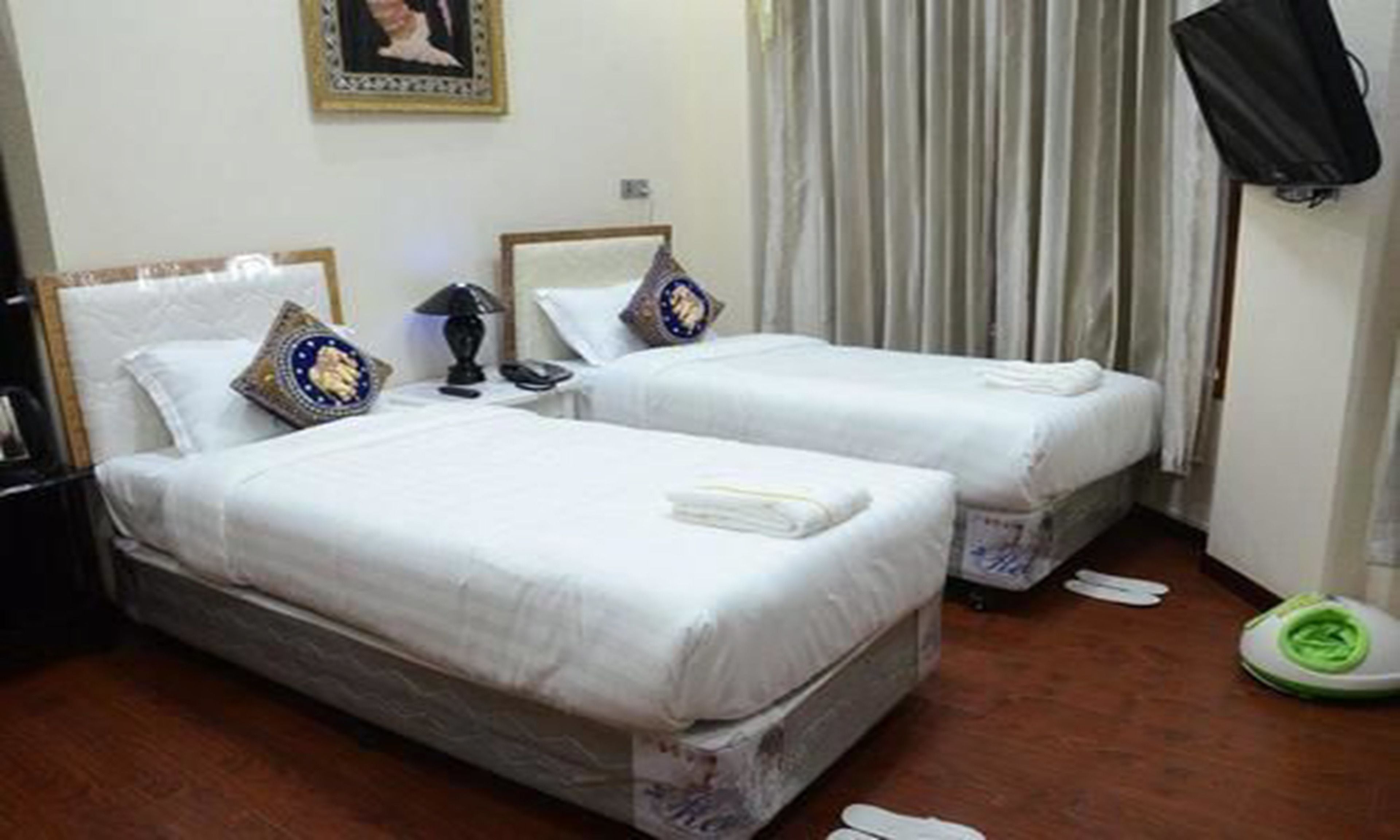 Habitación de hotel con dos camas individuales, una mesita de noche con lámpara, un cuadro enmarcado en la pared y una cortina.