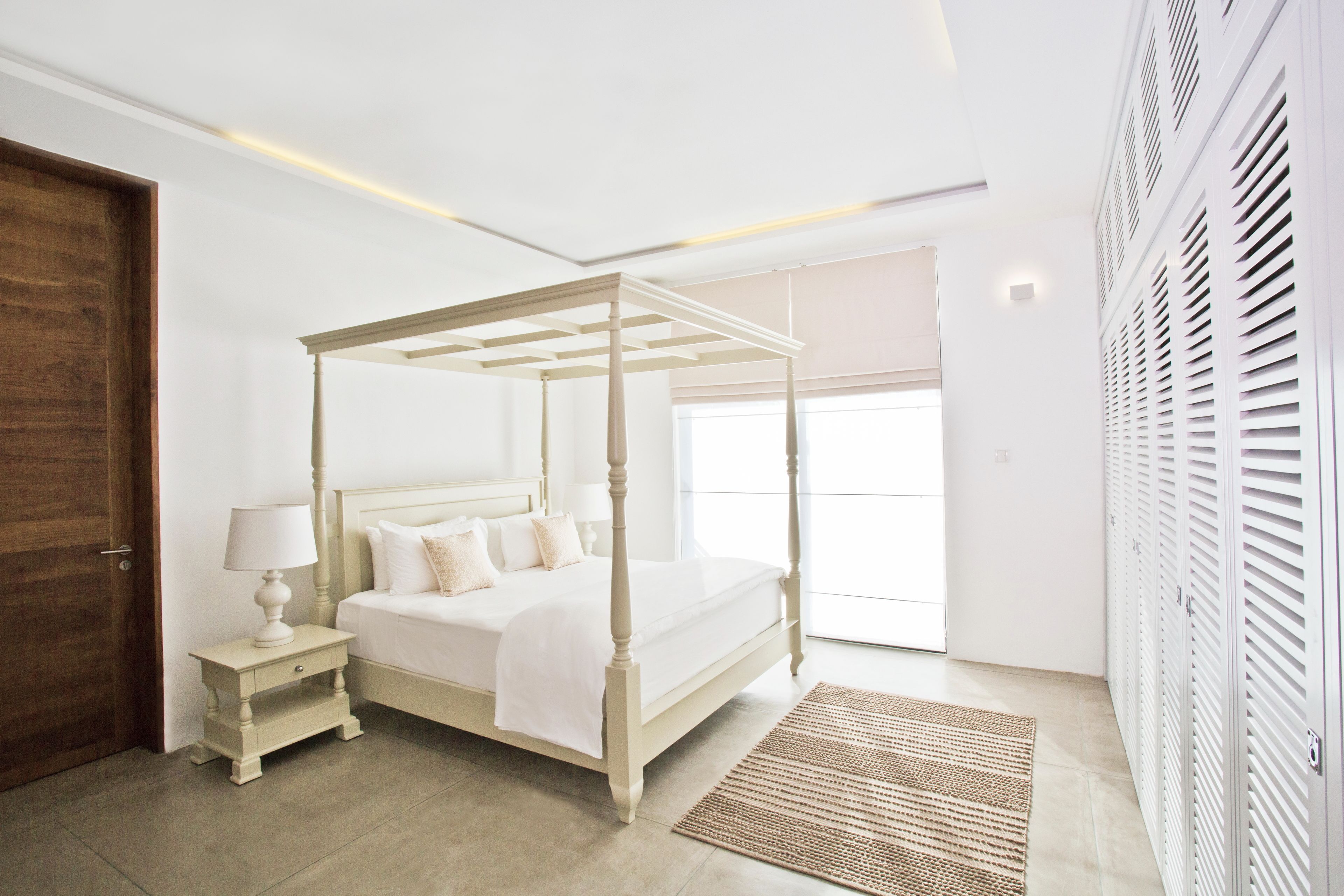 A four-poster bed with a nightstand, a lamp, and a woven rug in a room with white walls and a wooden door.