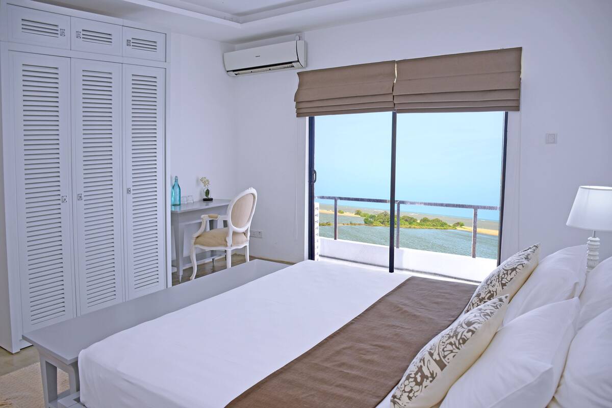 Une chambre avec un lit, un bureau et une chaise, une grande fenêtre donnant sur la mer et une armoire.
