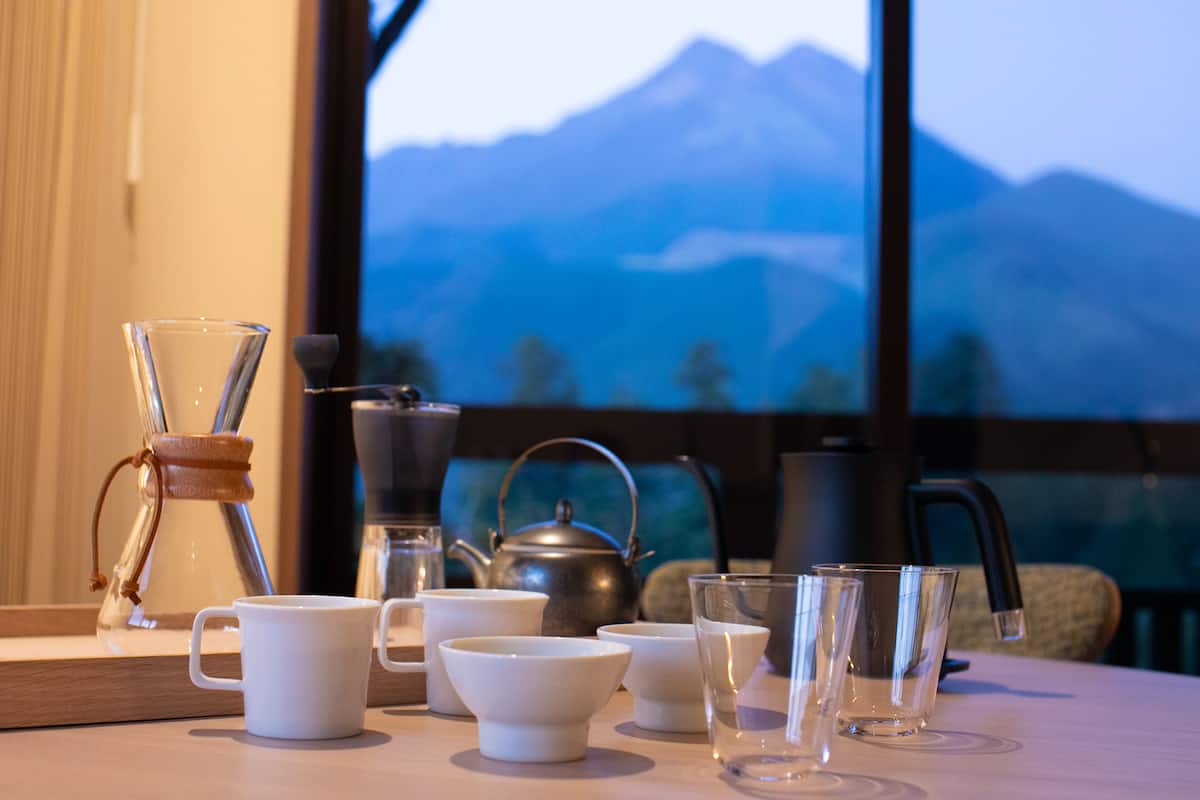 Ein Tisch mit verschiedenen Teetassen, Kaffeetassen und Gläsern, einer Teekanne und einer Kaffeemaschine, im Hintergrund ist ein Blick auf die Berge durch ein Fenster zu sehen.