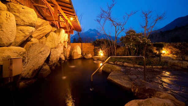 Uma onsen japonesa tradicional com estrutura de madeira, pedras grandes e um caminho de pedra que leva a uma nascente termal.