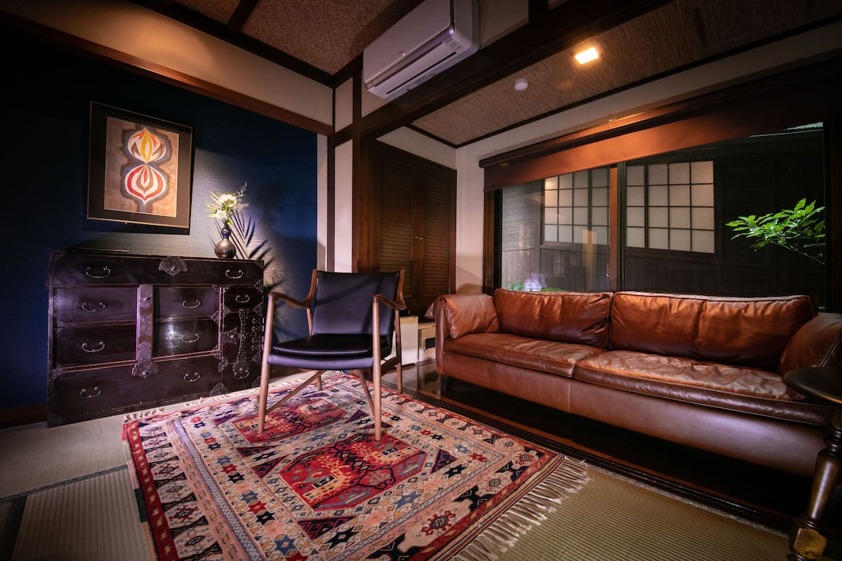 Ein traditionelles japanisches Wohnzimmer mit einer Ledersofa, einem dunklen Holzschrank, einem gemusterten Teppich und einer Wand-Klimaanlage.