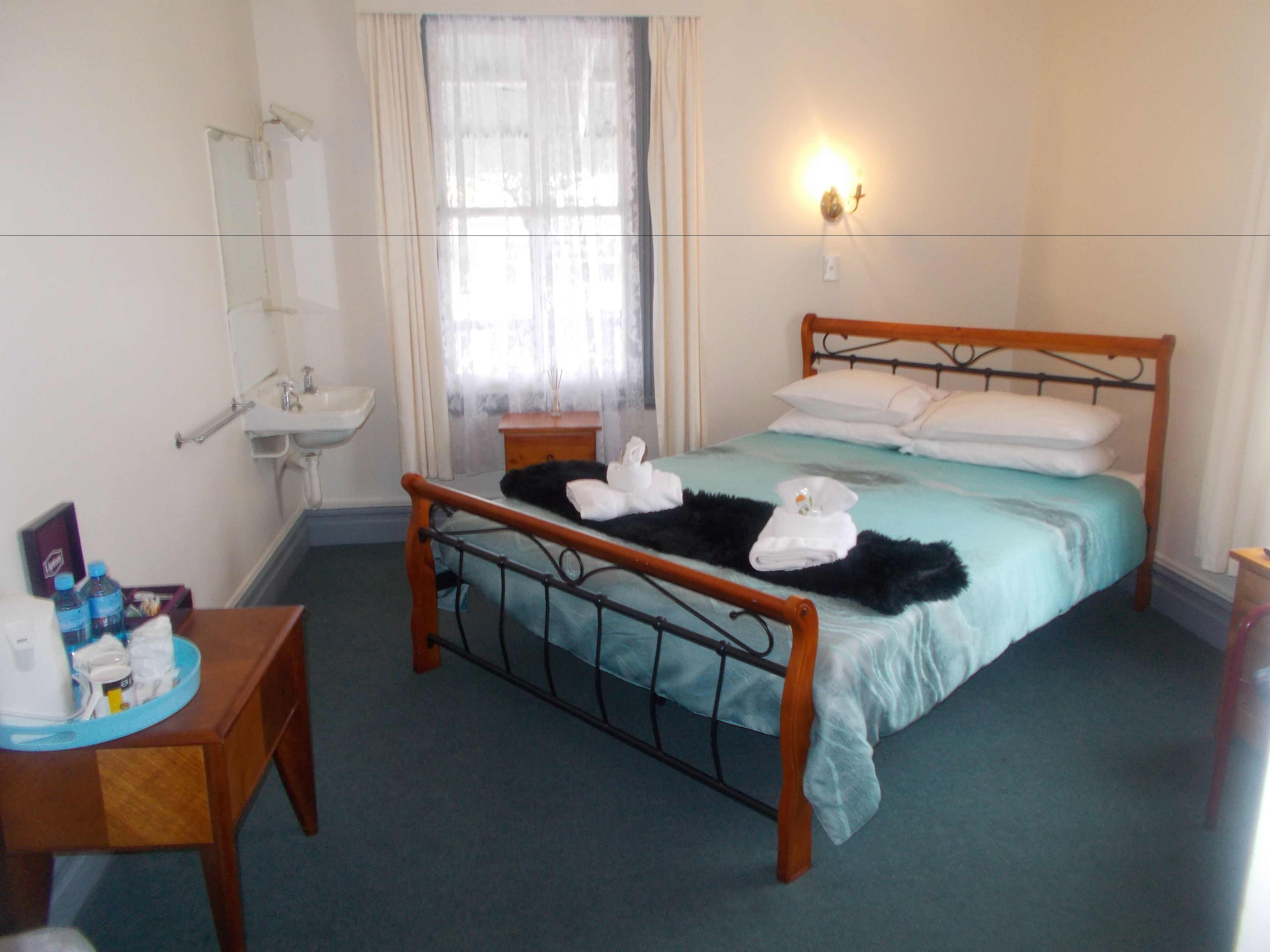 A hotel room with a bed, a bedside table, a nightstand, a window with curtains, a wall-mounted light, and a bathroom with a sink and mirror.