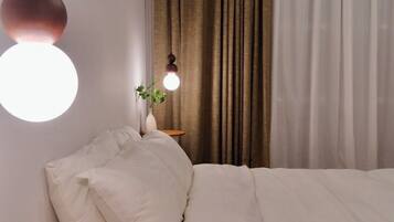 Quarto com cama, mesa de cabeceira com abajur, planta e janela com cortinas.