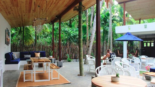 Un área de estar al aire libre con mesas y sillas, una estructura de madera cubierta y abundante vegetación al fondo.