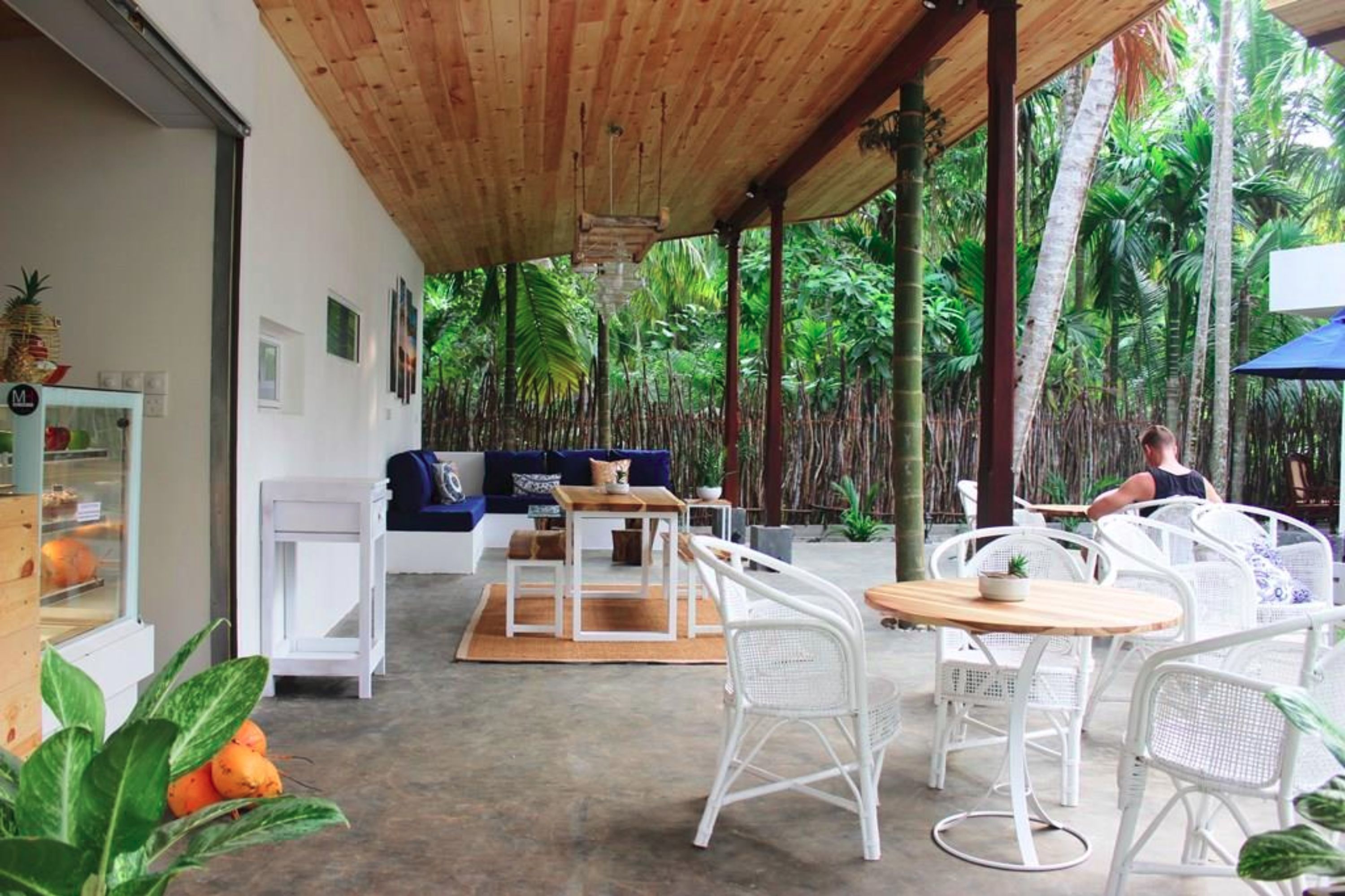 Un espace extérieur aménagé, abrité et doté de mobilier en osier blanc, d’un plafond en bois et offrant une vue sur une végétation luxuriante.