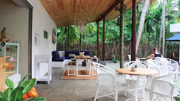 A covered outdoor seating area with white wicker furniture, a wooden ceiling, and a view of lush greenery.