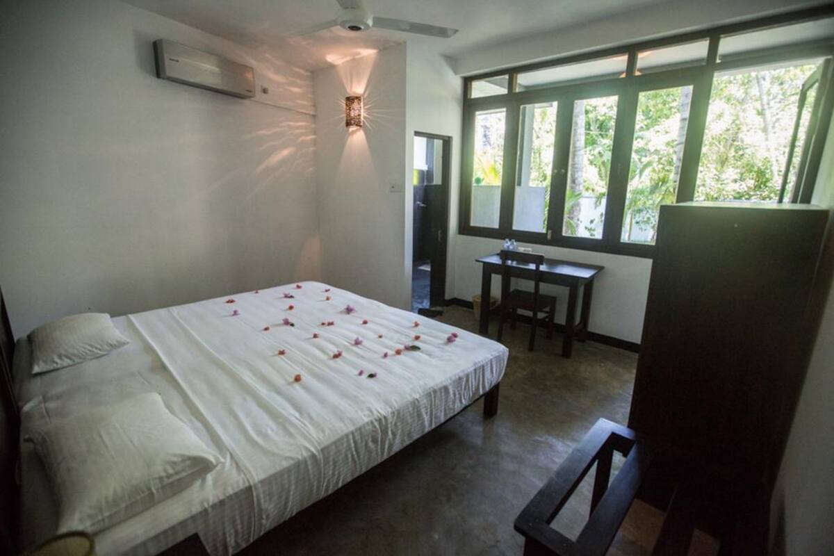 A bedroom with a bed, a desk, and a chair. There is a window with a view of greenery outside.