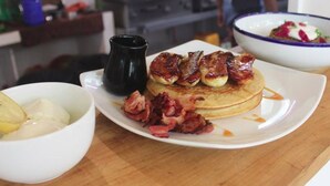 Un plato de desayuno con tortitas, tocino y un bol de fruta.