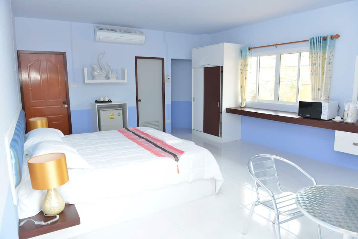 A hotel room with a bed, a desk with a computer, a chair, a window with curtains, and a small kitchenette.