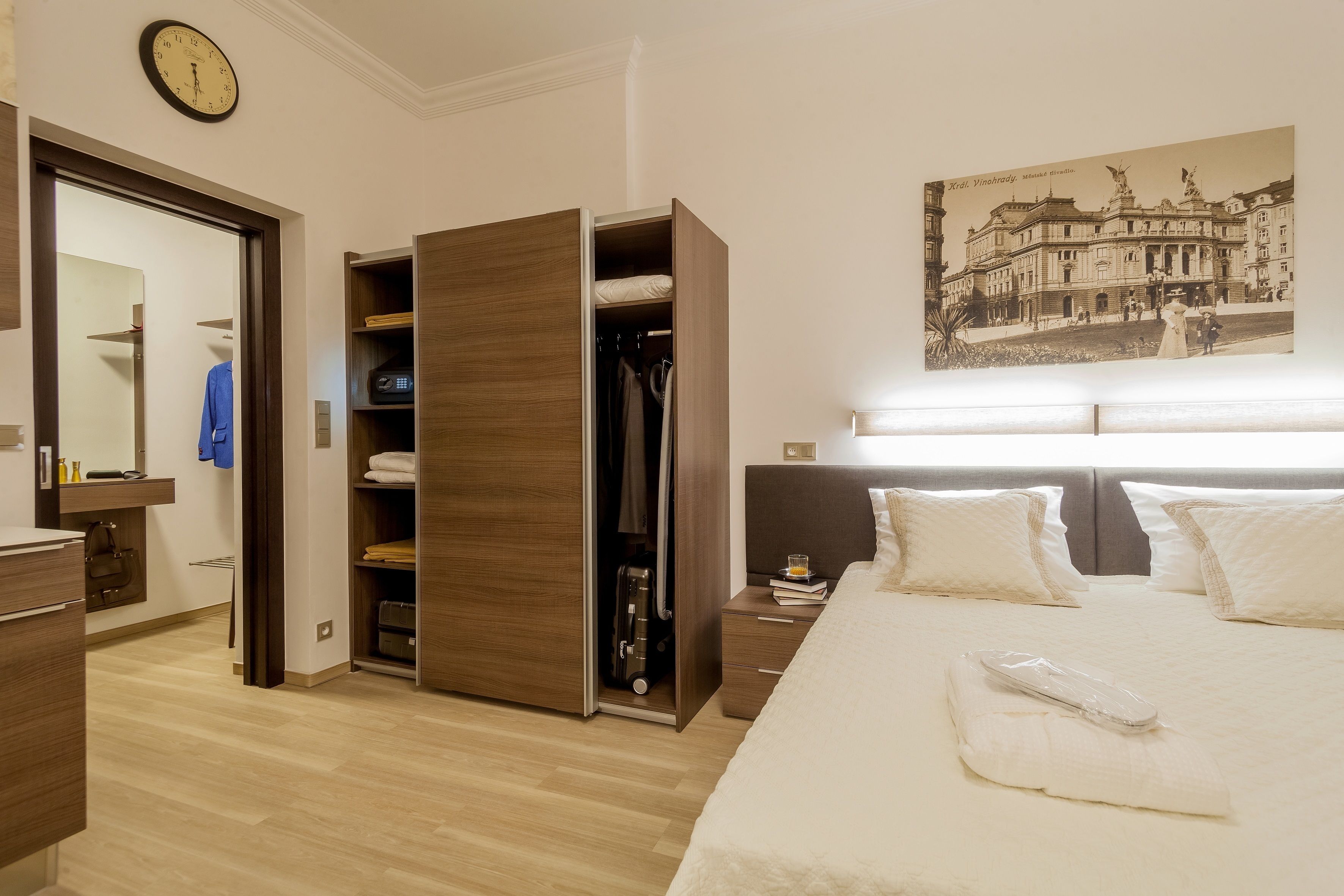 A hotel room with a bed, a wardrobe, a nightstand, a clock, and a painting on the wall.