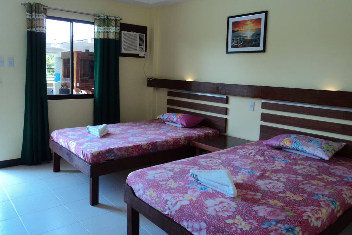 A room with two beds, each with floral bedding and pillows, a window with curtains, an air conditioning unit, and a framed picture on the wall.