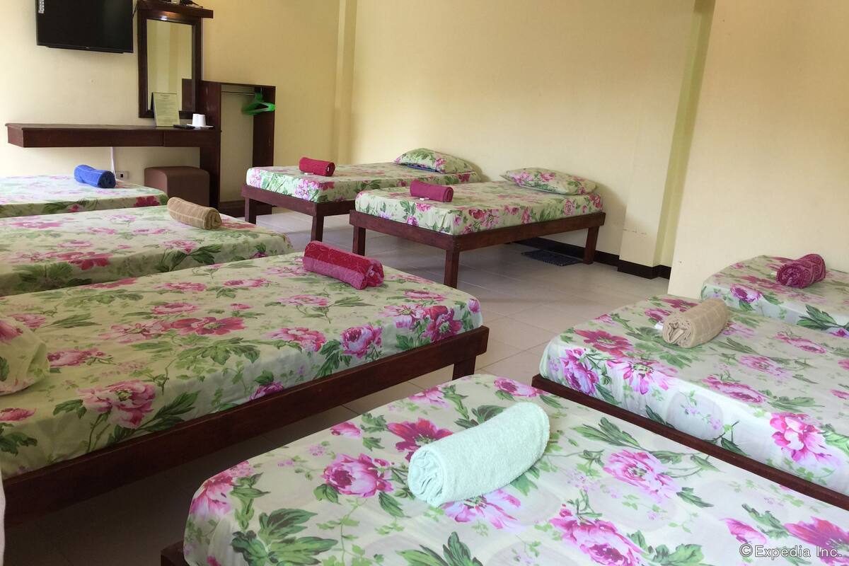 A hotel room with four single beds, each with floral bedding and a rolled towel. There is a desk with a flat-screen TV and a mirror above it.