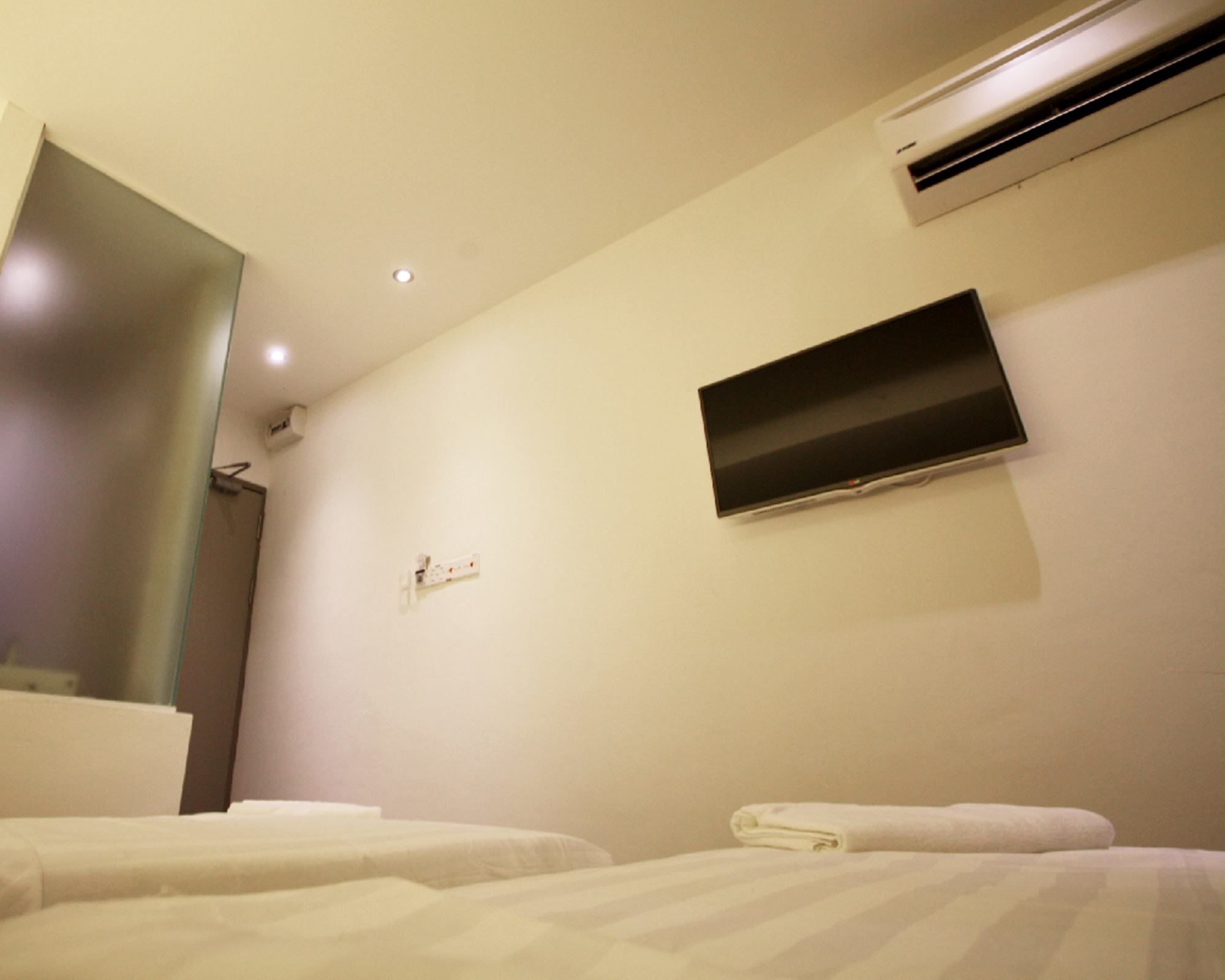 A hotel room with two beds, a flat-screen TV mounted on the wall, an air conditioning unit, and a small bedside table with a lamp.