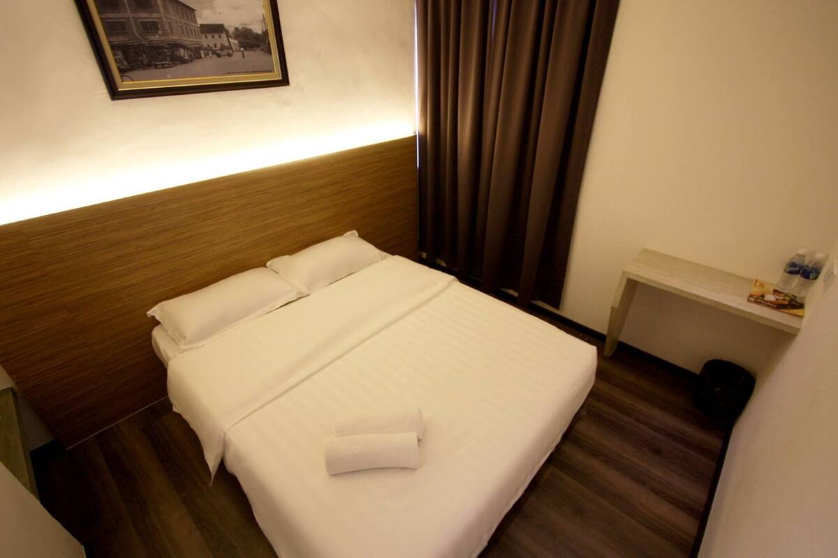 A hotel room with a neatly made bed, a small bedside table with a bottle, and a framed picture on the wall.