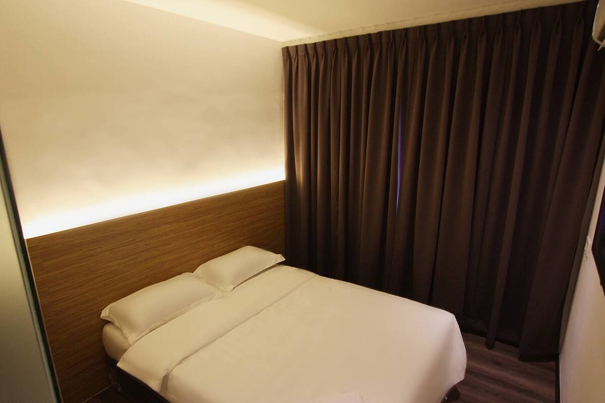 A hotel room with a neatly made bed, a wooden headboard, and a dark curtain.
