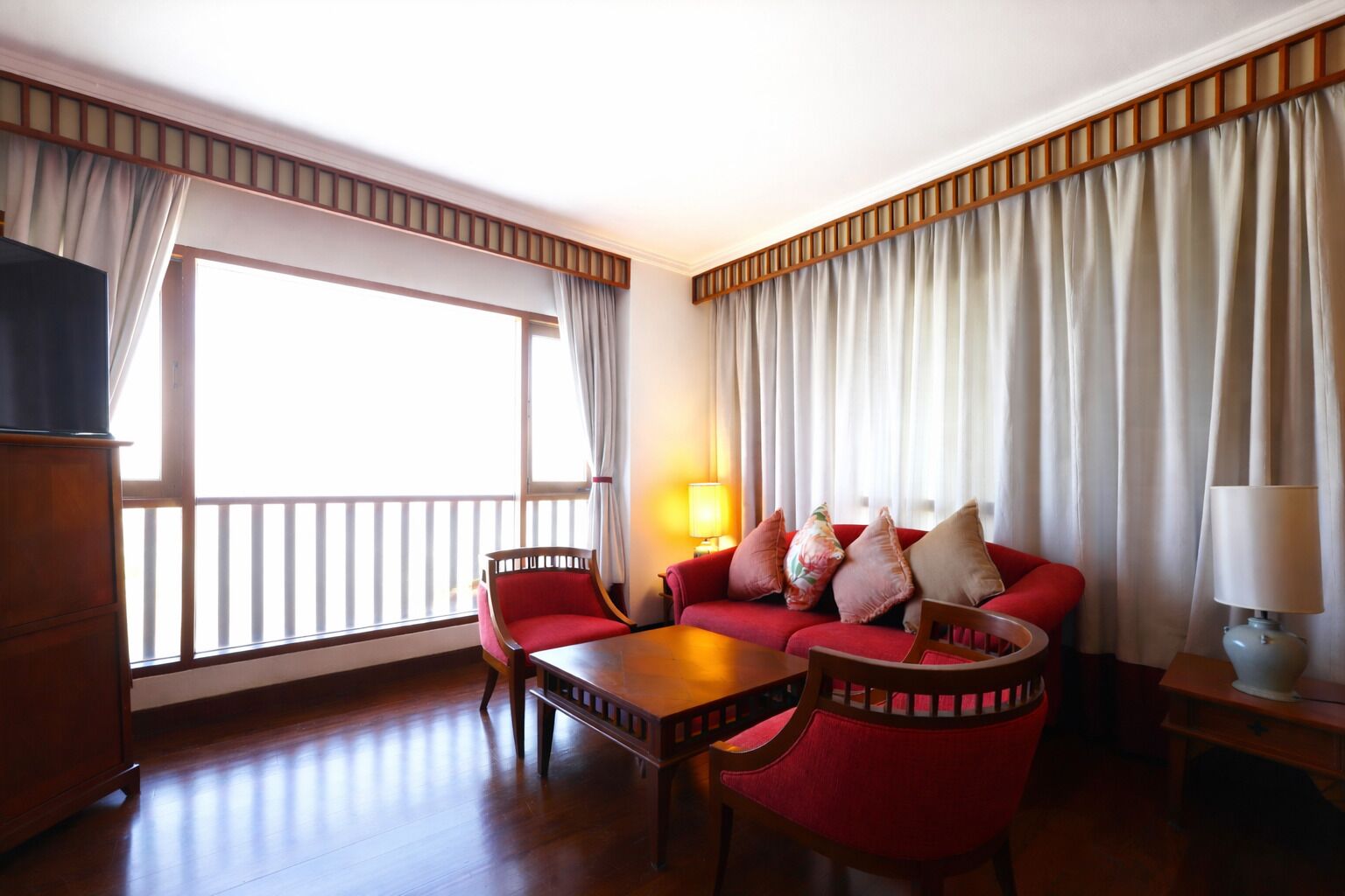 Habitación de hotel con sofá rojo, una mesa de centro de madera, televisor y un ventanal con cortinas.