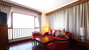 Habitación de hotel con sofá rojo, una mesa de centro de madera, televisor y un ventanal con cortinas.