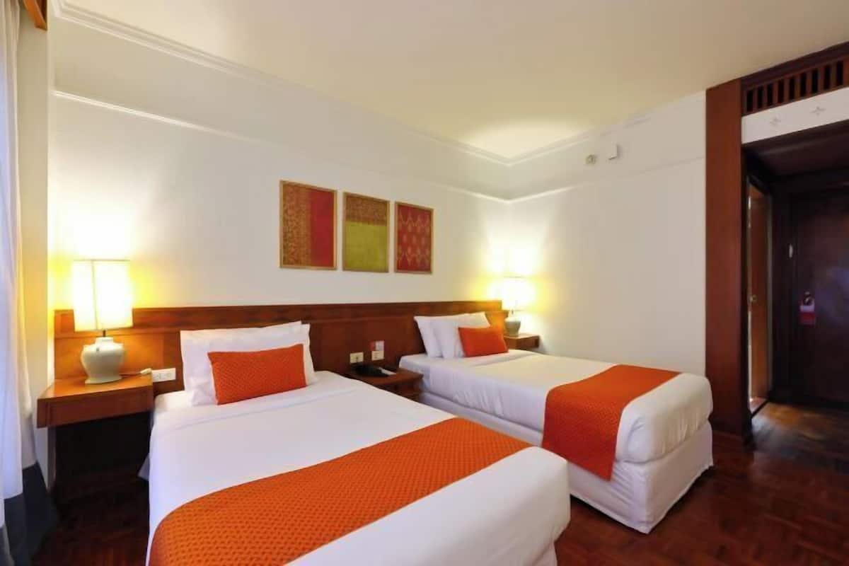 Habitación de hotel con dos camas, cada una con cubrecama blanco y naranja, cabecero de madera y mesita de noche con lámpara.