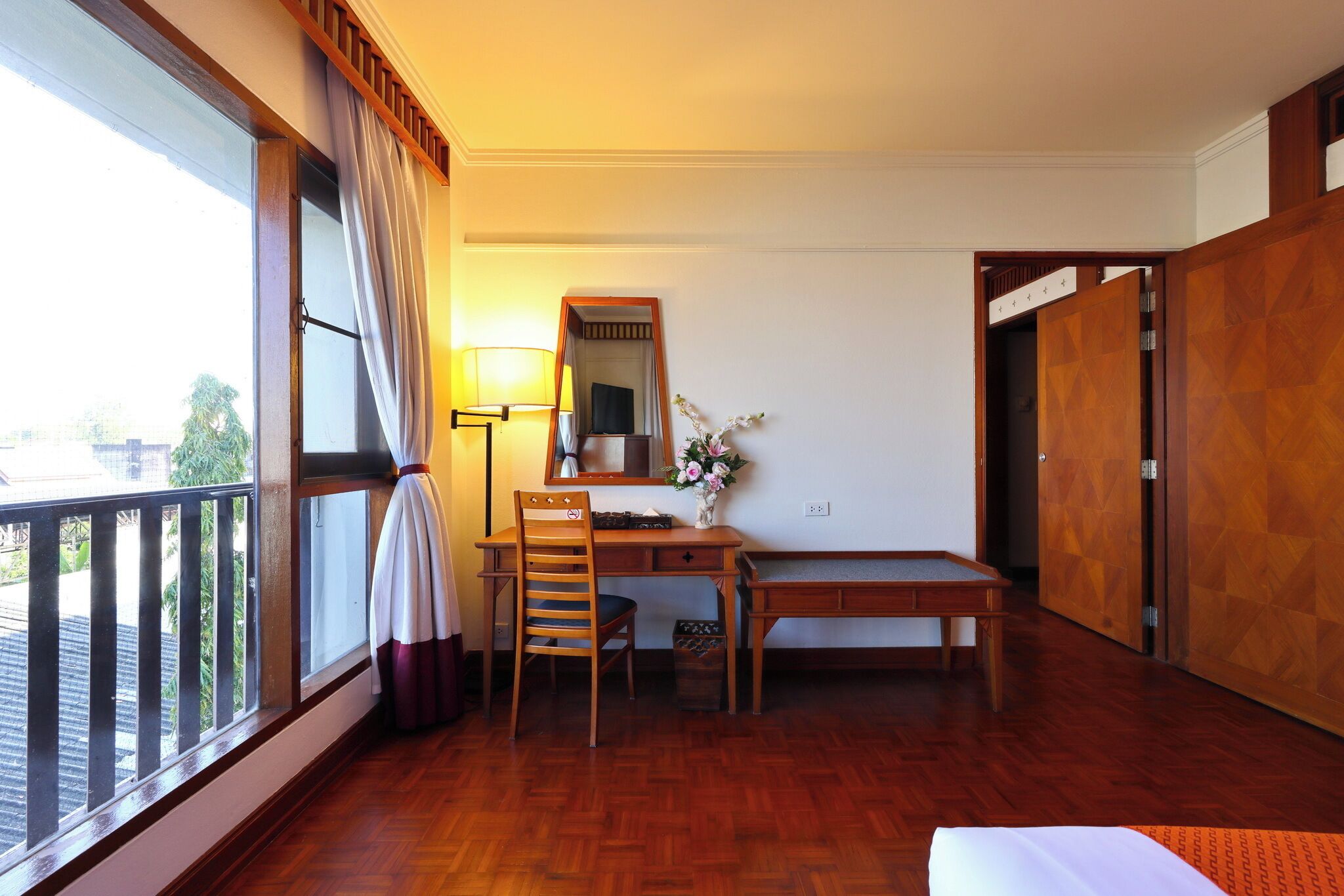 Habitación de hotel con un escritorio de madera, una silla y una mesita. Hay una ventana con cortinas, un espejo y una lámpara.