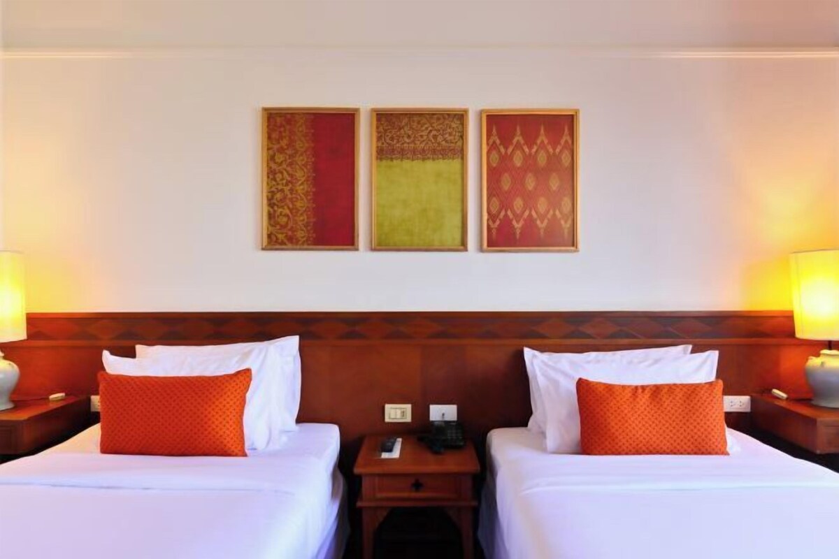 A hotel room with two beds, a wooden headboard, a nightstand with a lamp, and three framed artworks on the wall.