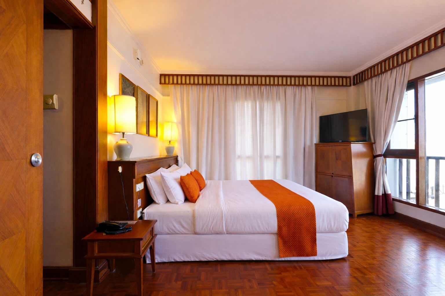 A hotel room with a bed, bedside lamps, a TV, a wooden wardrobe, and a window with curtains.