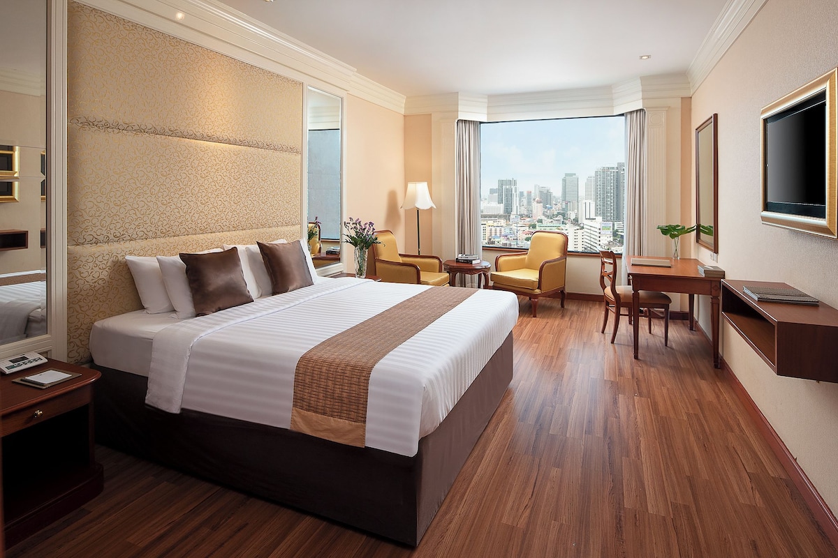 A hotel room with a large bed, a desk, a chair, a TV, and a view of the city.