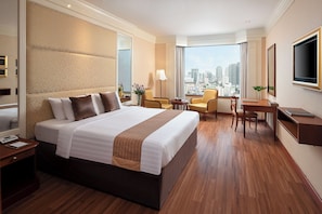 A hotel room with a large bed, a desk, a chair, a TV, and a view of the city. - Emerald Hotel Bangkok (Bangkok)