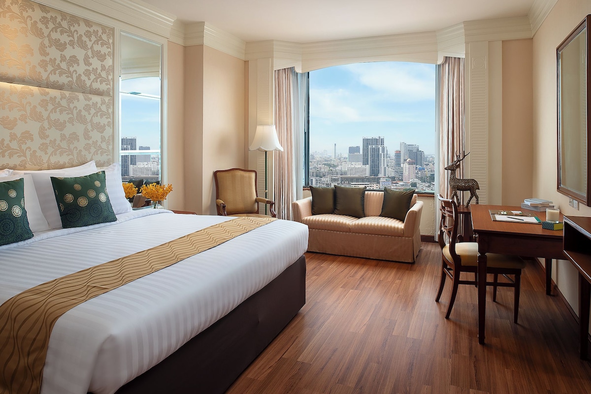 A hotel room with a large bed, a sofa, a dining table, and a view of the city.