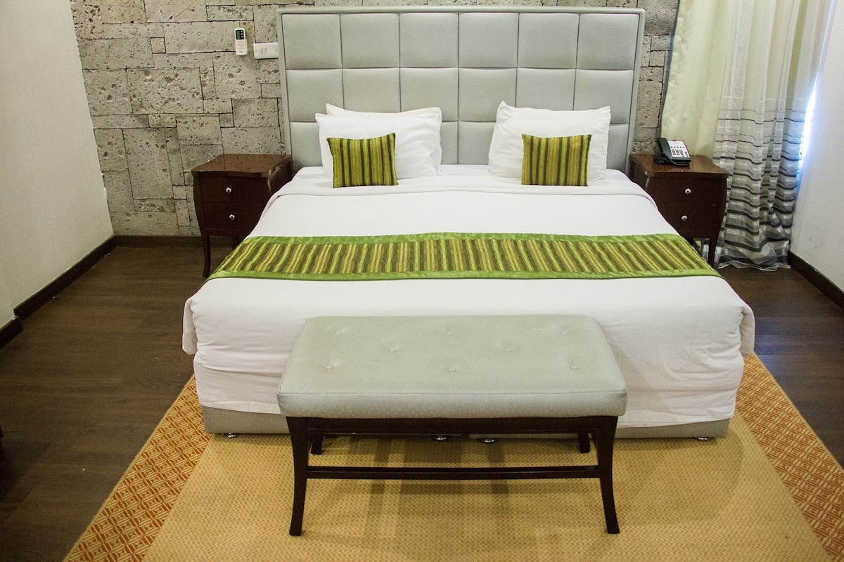 Una cama bien tendida con cabecera gris, dos mesitas de noche, un teléfono y un banco en una habitación de hotel.