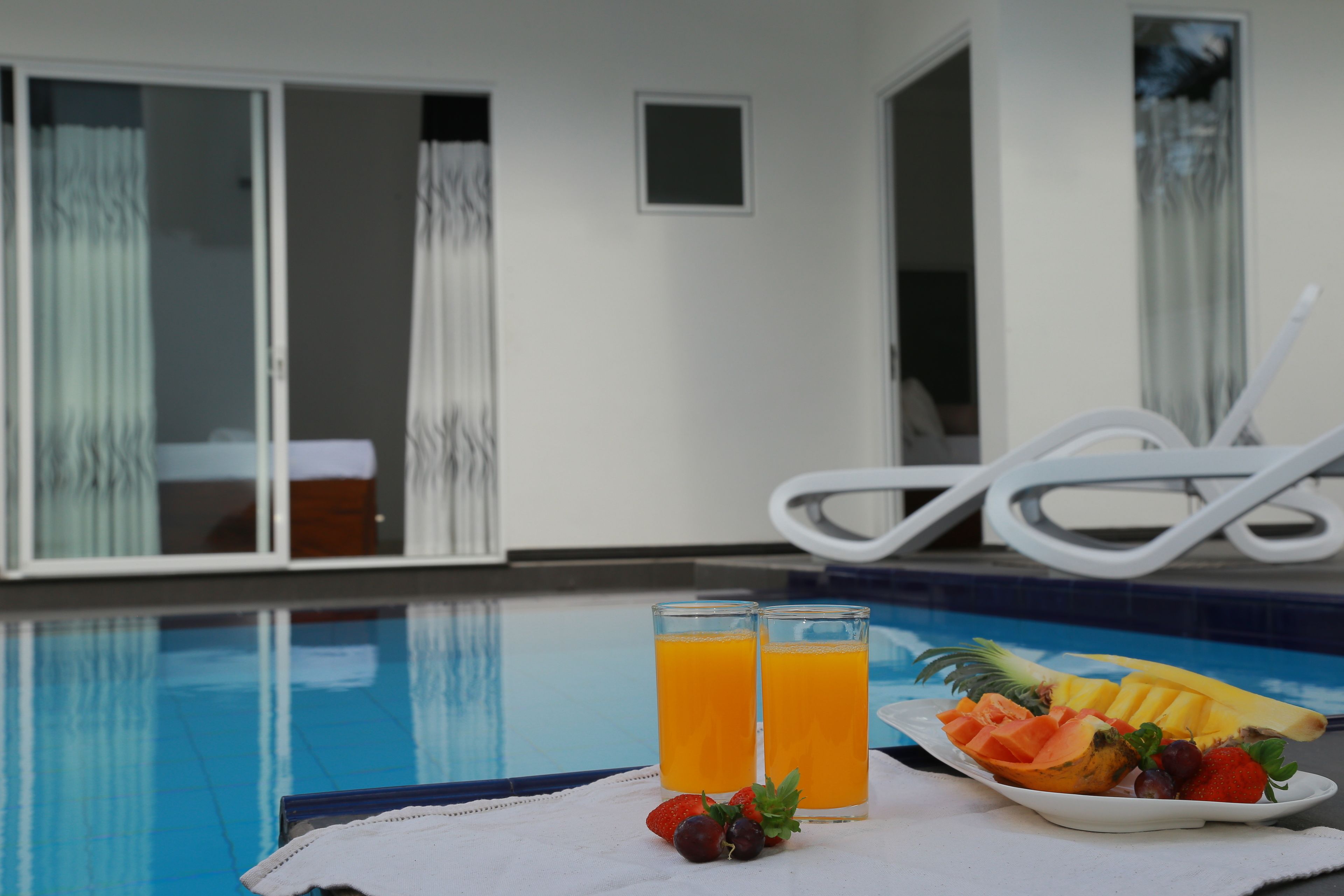A poolside setting with a table holding two glasses of orange juice, a plate of fruit, and a towel.