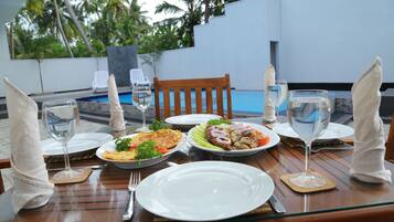 Mesa de comedor con platos, cubiertos y vasos, con vistas a una piscina rodeada de palmeras.