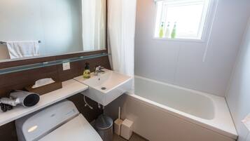 A modern bathroom with a white sink, mirror, and bathtub. There is a window with bottles on the windowsill and a towel hanging on the wall.